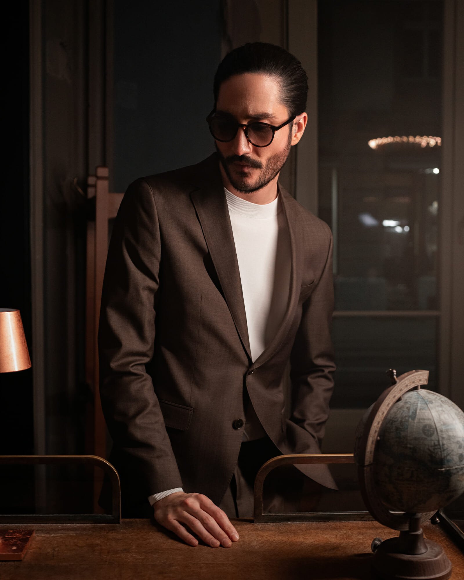 Asian dark haired man. He is standing behind a wooden table, wearing an elegant brown suit and black sunglasses. He looks down to the right