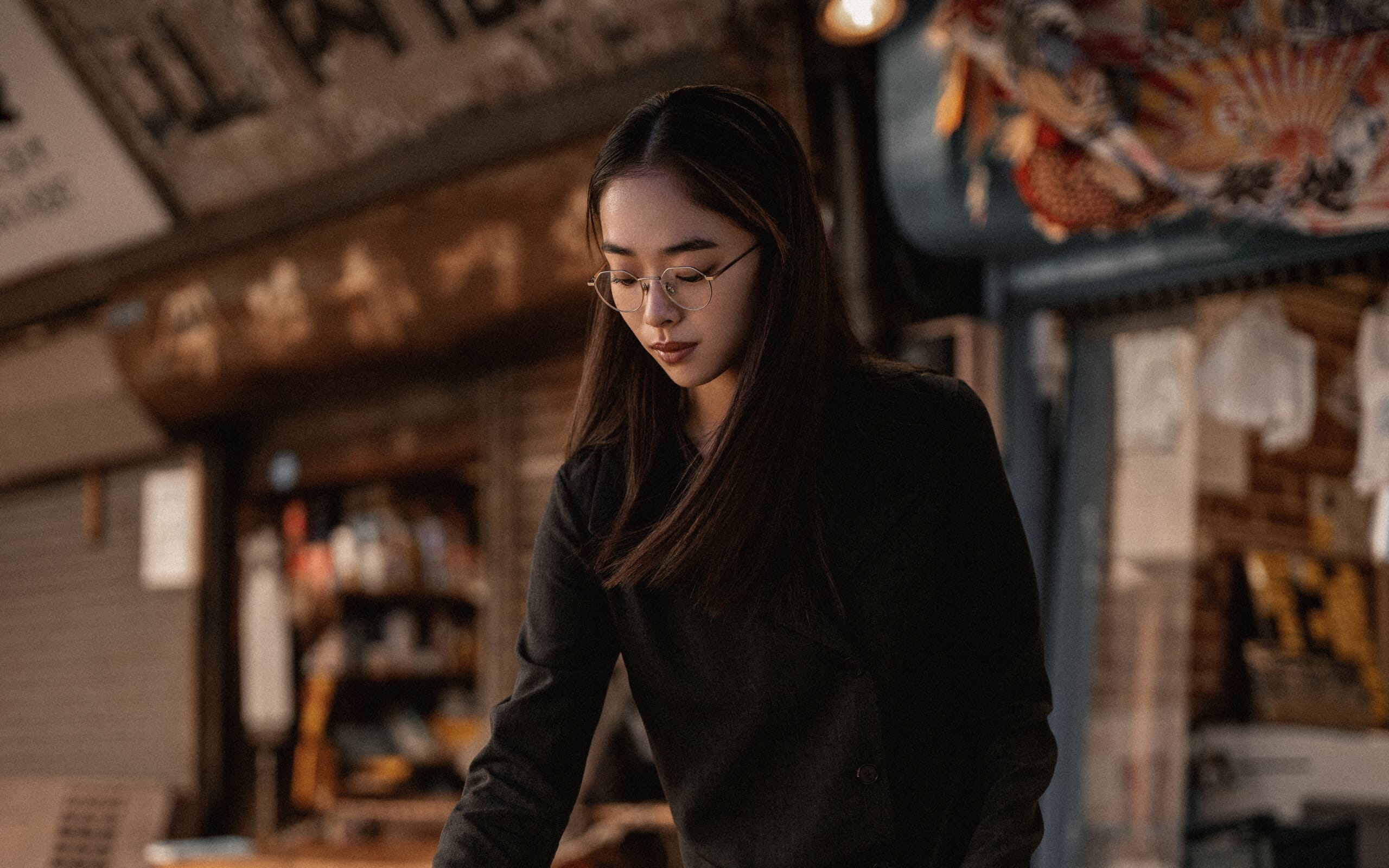 A woman wearing Nirvan Javan's minimalist Tokyo optical frames thoughtfully examines items at a traditional Japanese market, embodying the understated elegance and precision of the TOKYO Collection.