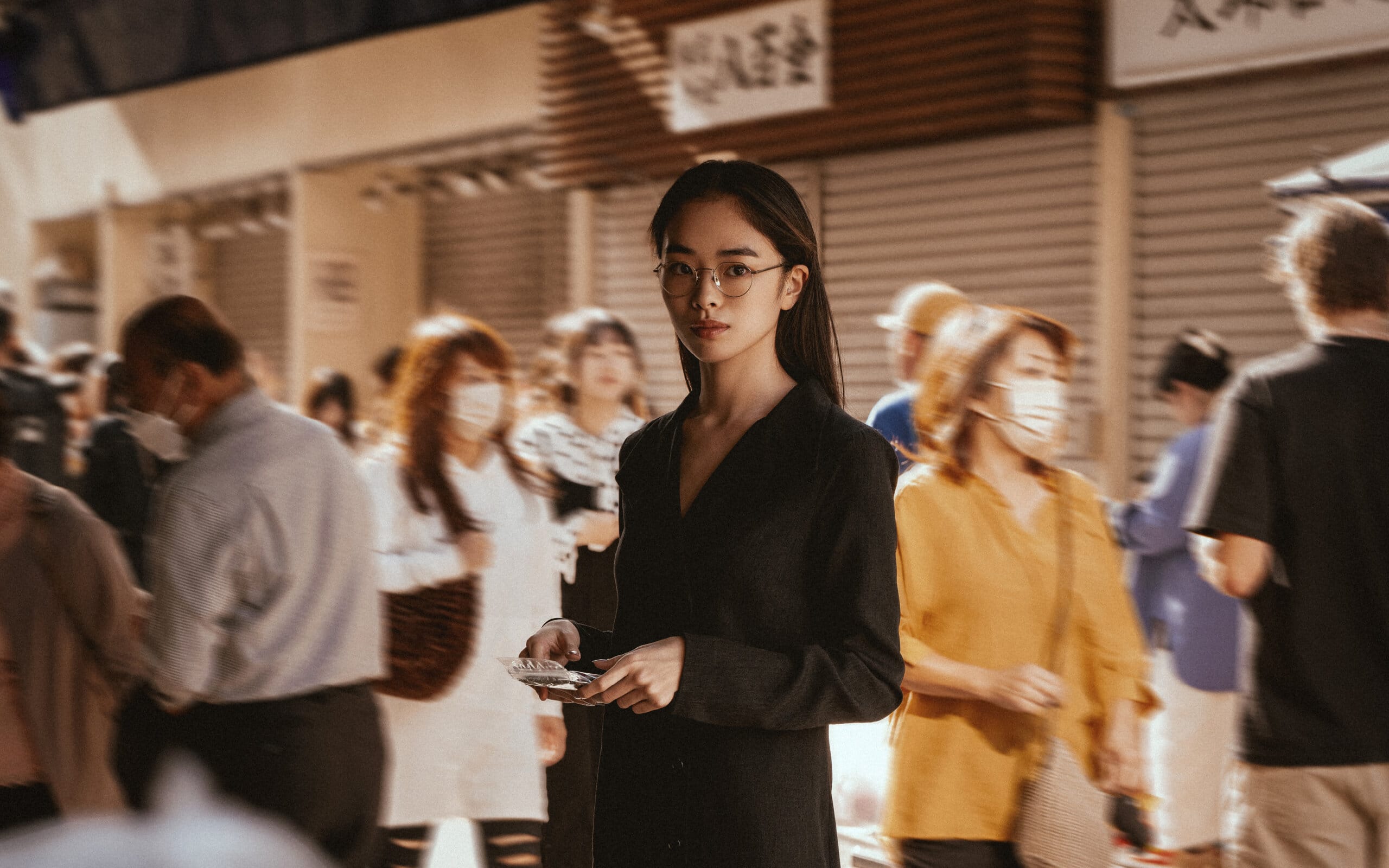 A woman wearing Nirvan Javan's Tokyo optical frames stands in a busy Japanese street market, exuding calm and elegance amidst the bustling crowd, reflecting the refined and thoughtful design of the TOKYO Collection.